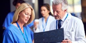 Two Doctors In White Coat And Scrubs Discussing Patient Scan In Busy Hospital